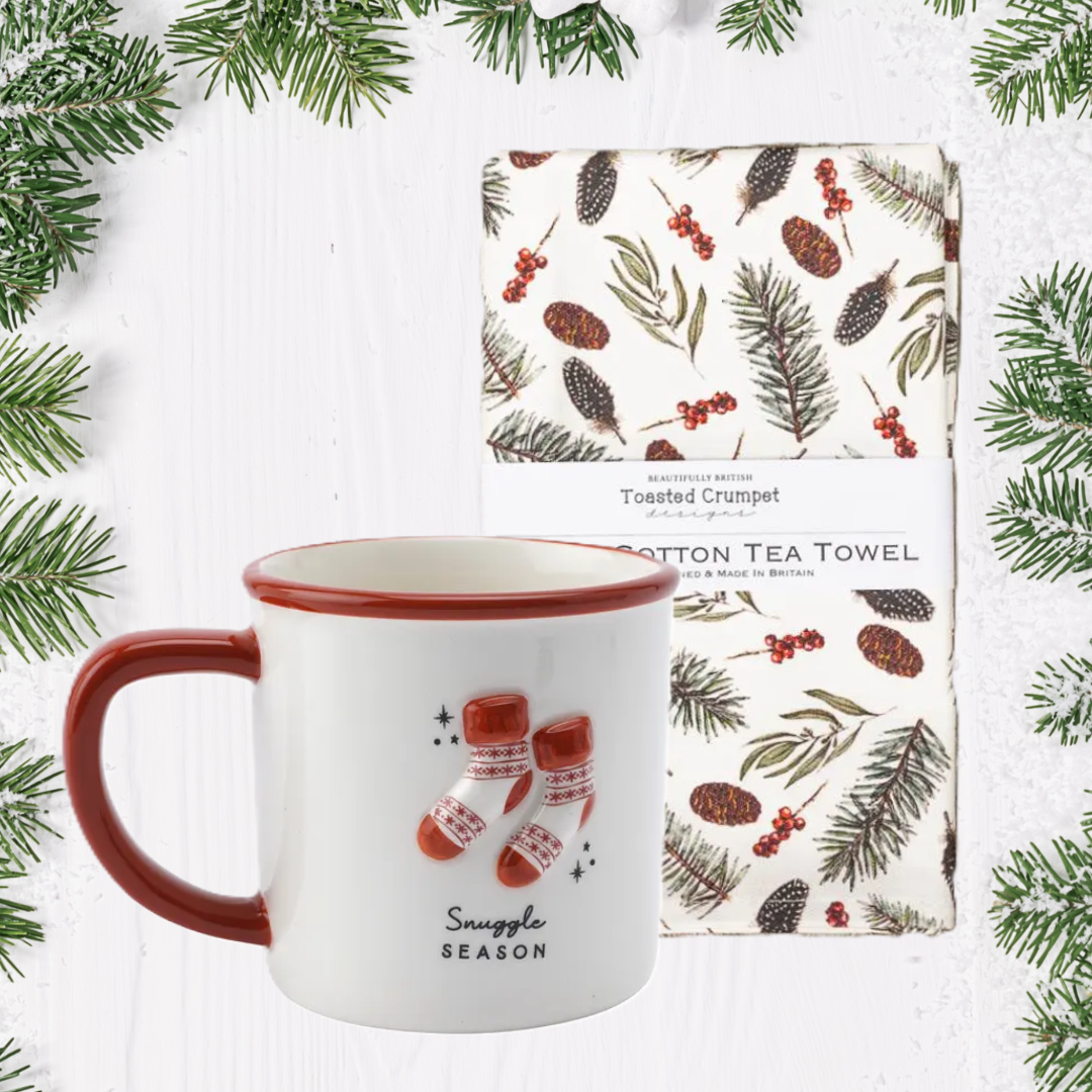 White mug with red interior and handle, featuring Christmas-themed design, next to a patterned tea towel on a white background with greenery.