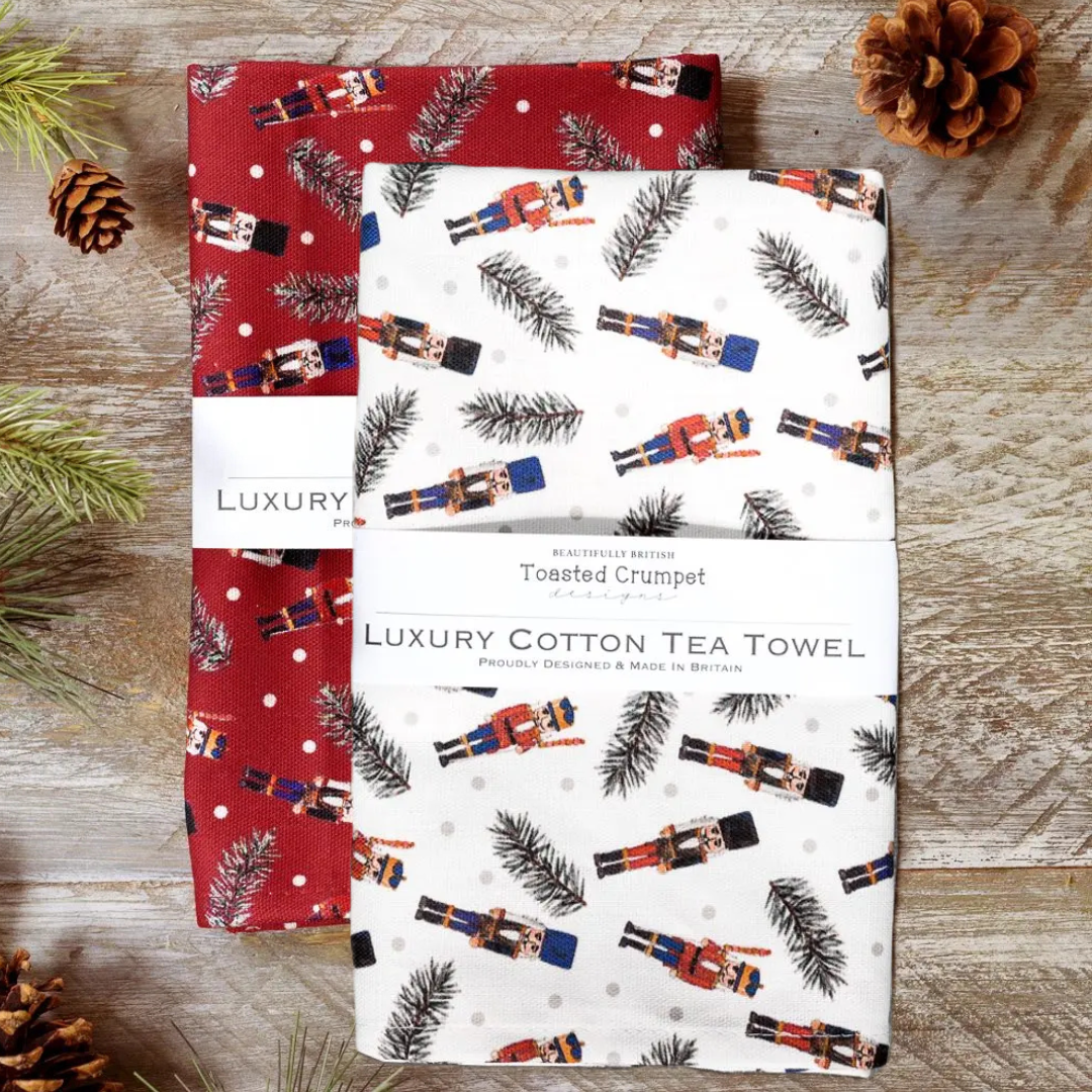 Two tea towels red and white with festive designs on a wooden surface with pine cones and branches.