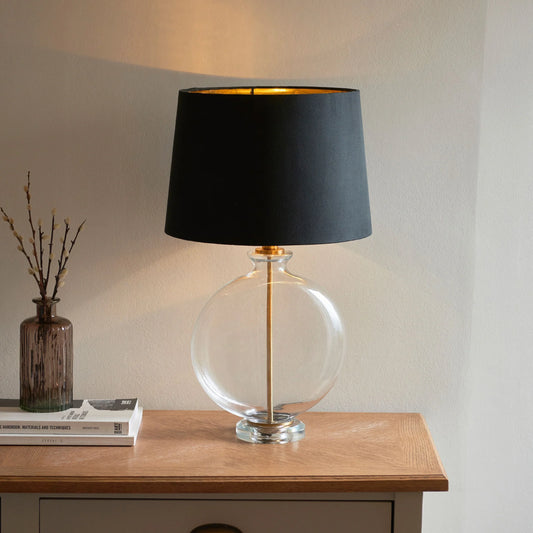 Table lamp with black shade and clear glass base on a wooden surface.