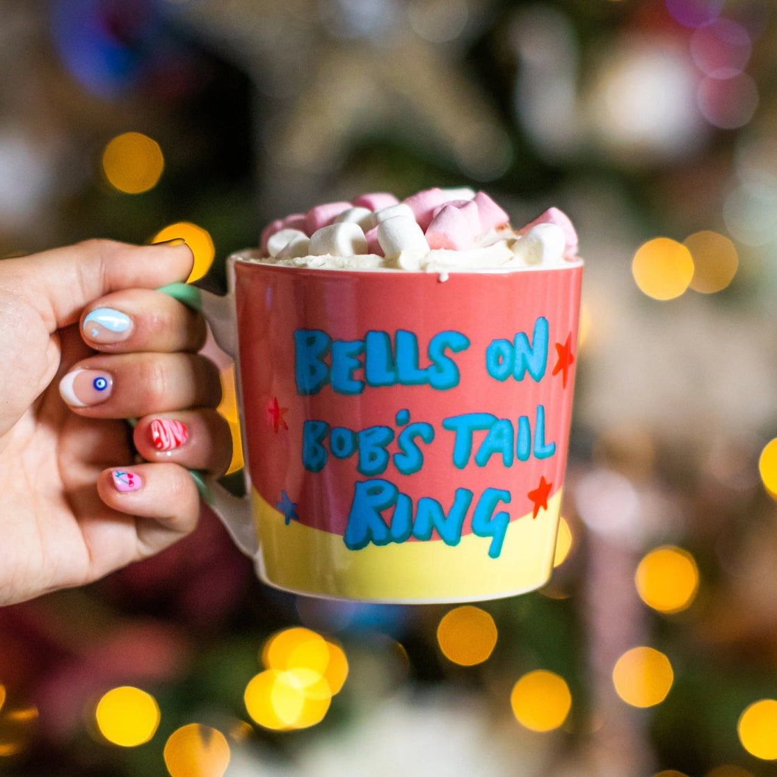 Hand holding a mug with 'Bells on Bob's Tail Ring' text against a festive background