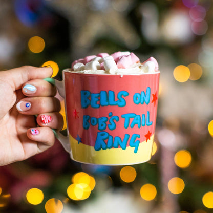 Hand holding a mug with 'Bells on Bob's Tail Ring' text against a festive background