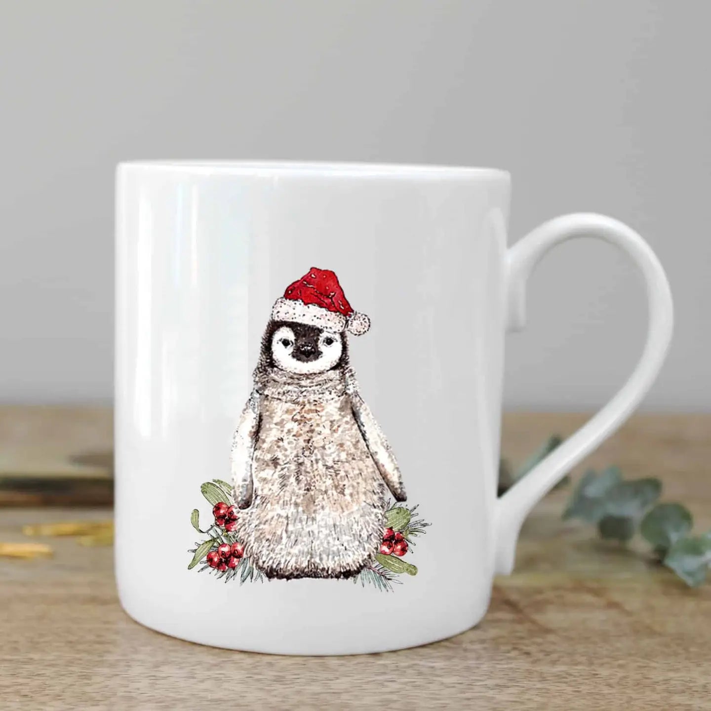 White mug with a penguin wearing a Santa hat and holly leaves on a wooden surface.
