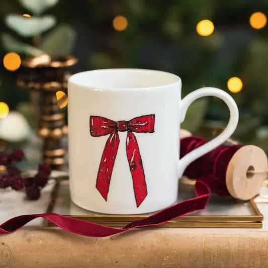 White mug with red bow design on a wooden surface with Christmas decorations in the background