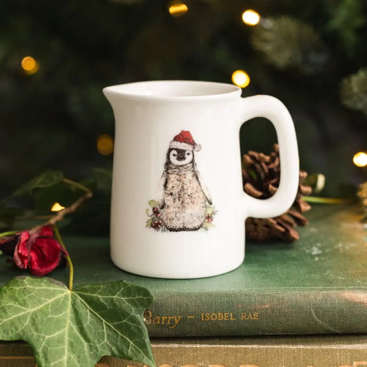 White mug with a penguin design wearing a Santa hat on a green surface with festive decorations.