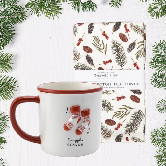 White mug with red interior and handle, featuring Christmas-themed design, next to a patterned tea towel on a white background with greenery.