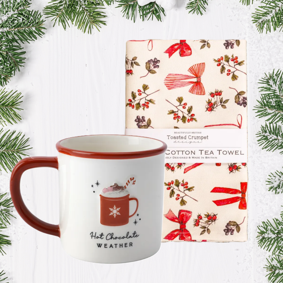 Mug with 'Hot Chocolate Weather' text and design, next to a cotton tea towel with festive pattern, surrounded by Christmas tree branches.