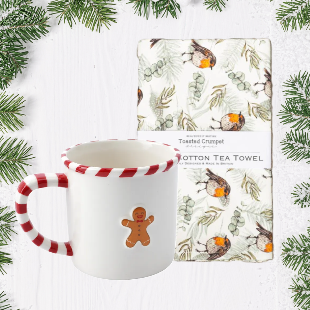 White mug with red and white handle and gingerbread man design, next to a cotton tea towel with nature-themed design on a white background with greenery.