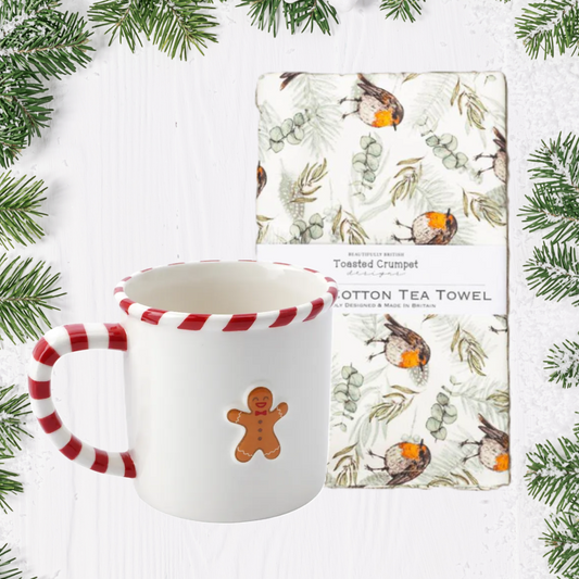 White mug with red and white handle and gingerbread man design, next to a cotton tea towel with nature-themed design on a white background with greenery.