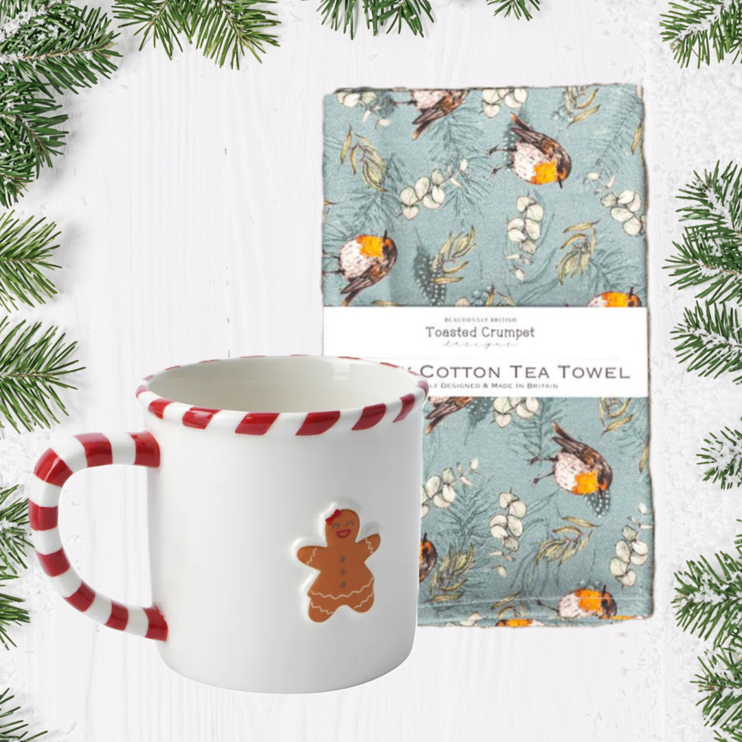 Ceramic mug with candy cane handle and gingerbread man design, accompanied by a cotton tea towel with nature-themed design, surrounded by Christmas tree branches.