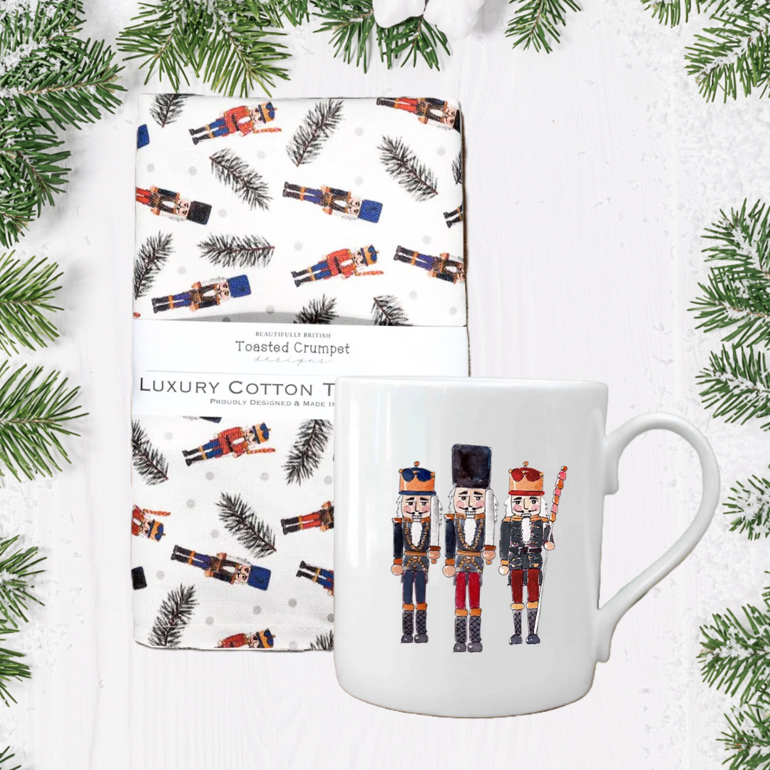 White mug with nutcrackers design on a white background with tea towel, and Christmas tree branches.