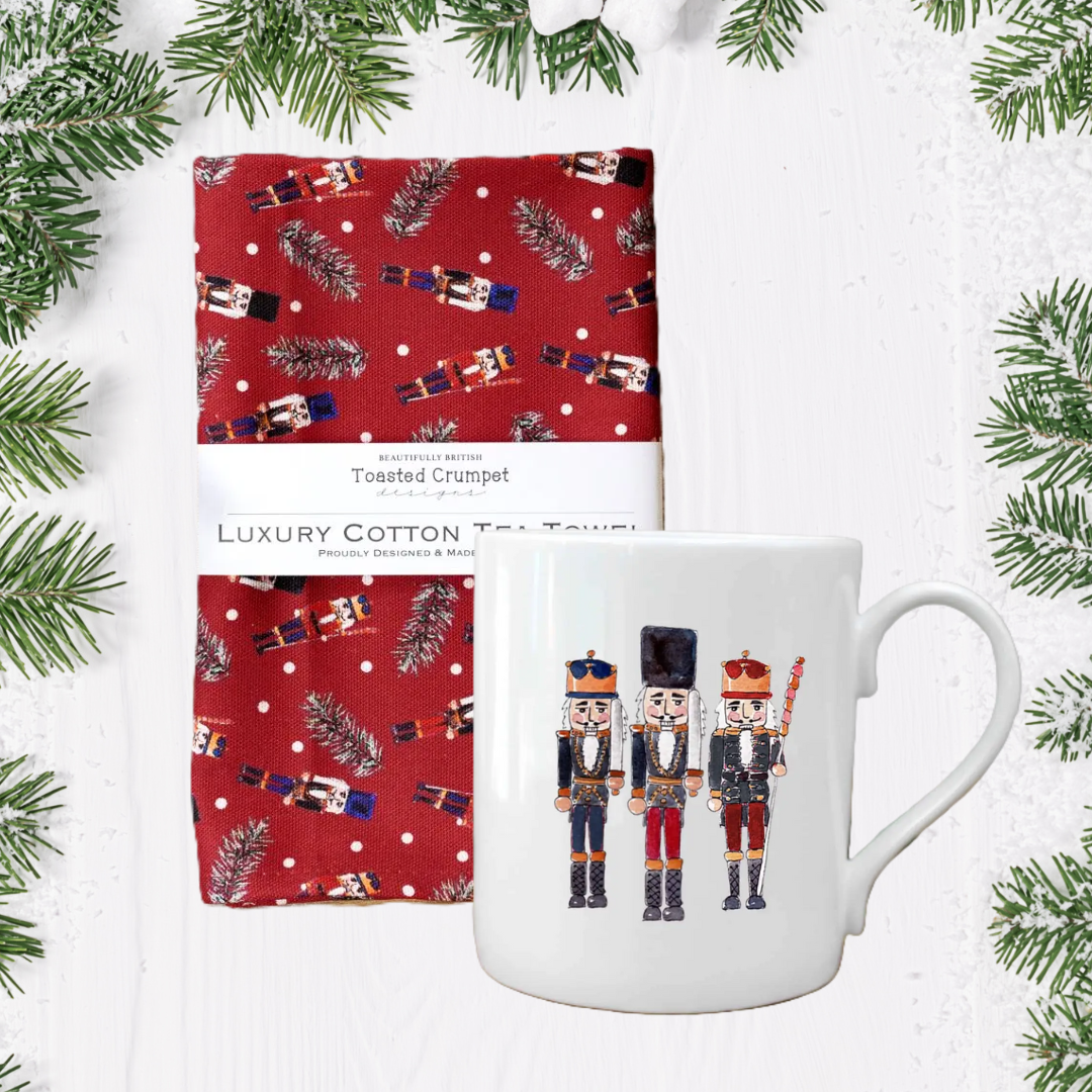 White mug with nutcrackers design and red patterned towel on a white background with Christmas tree branches.
