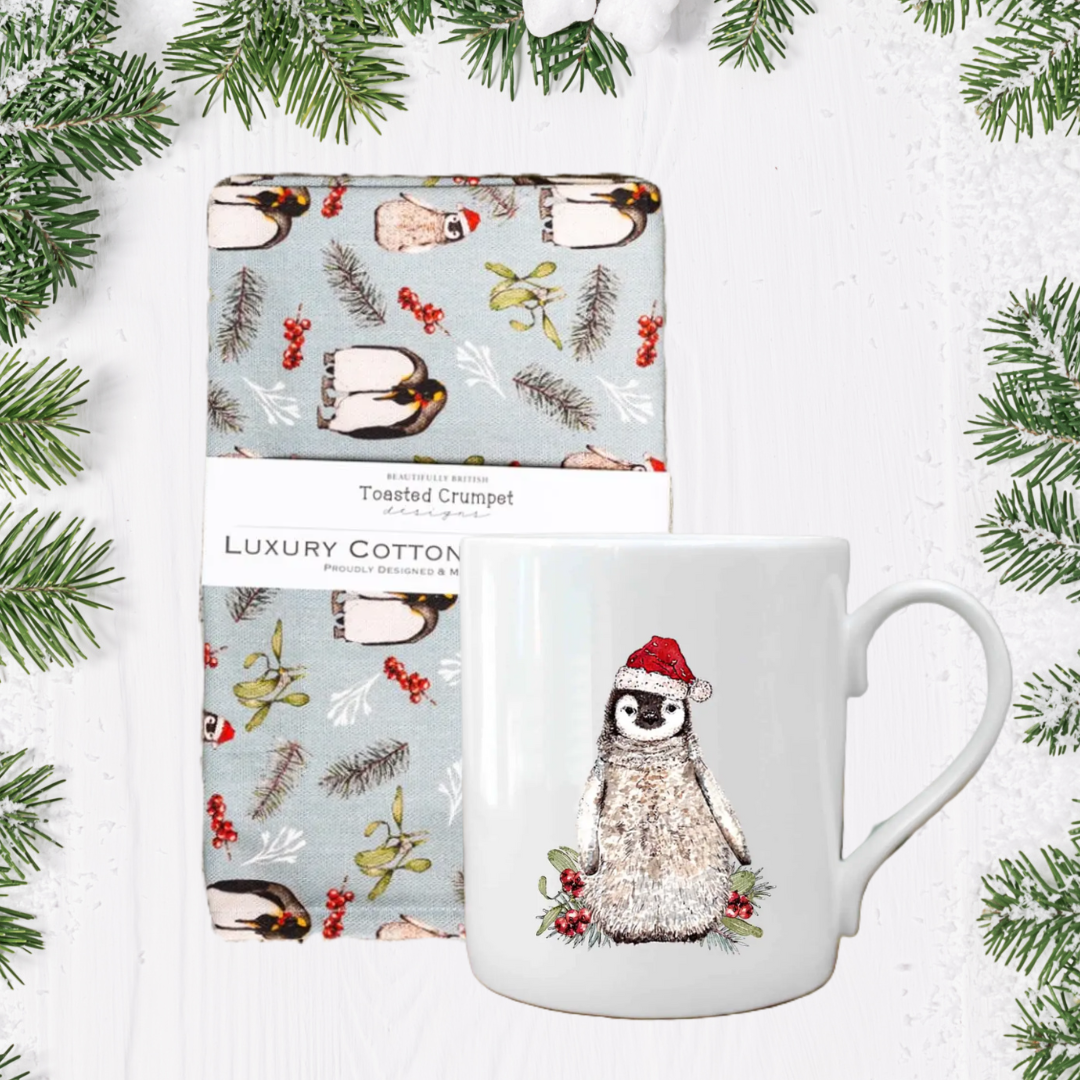 Mug with penguin design and patterned penguin tea towel on a white background with greenery.