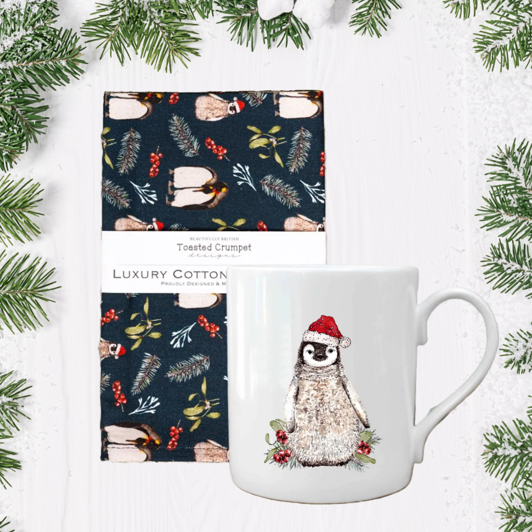 Mug with penguin design and tea towel with Christmas pattern on a white background with greenery.