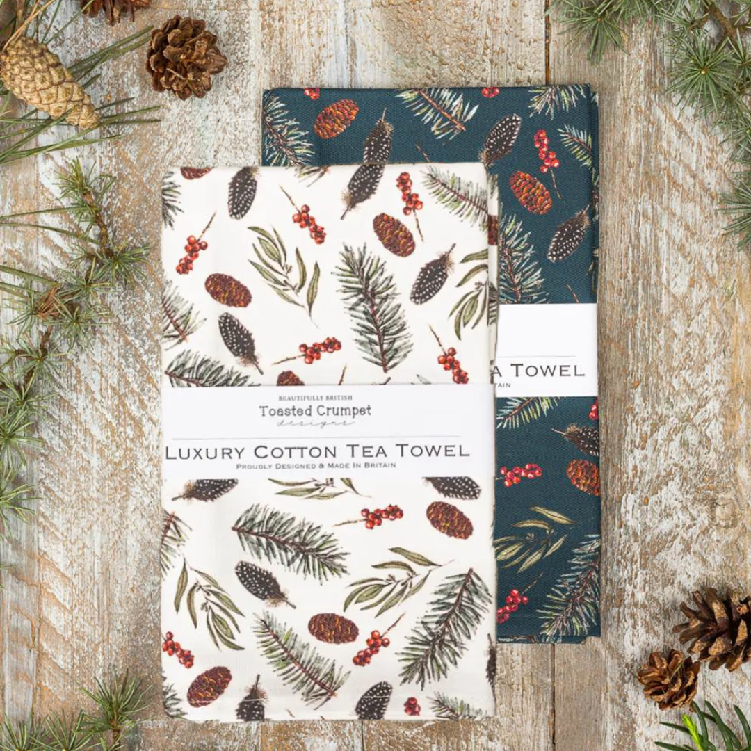 Two tea towels with nature-themed designs on a wooden surface with pinecones and branches.