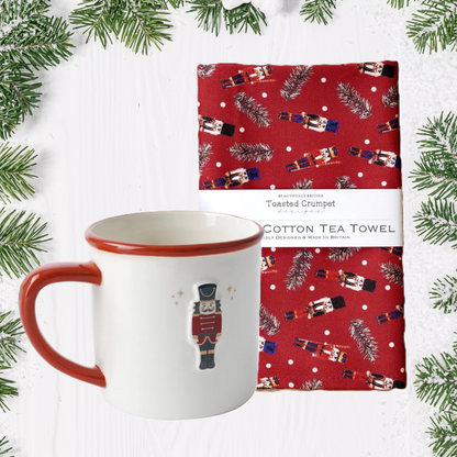 Ceramic mug with Nutcracker and red handle, next to a red cotton tea towel with Christmas design on a white background with greenery.