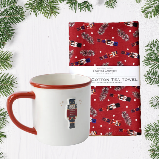 Ceramic mug with Nutcracker and red handle, next to a red cotton tea towel with Christmas design on a white background with greenery.