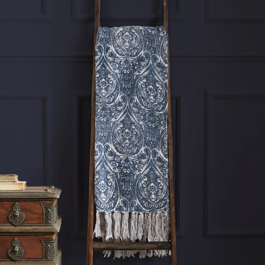 Decorative blue and white damask patterned throw blanket on a wooden ladder against a dark wall.