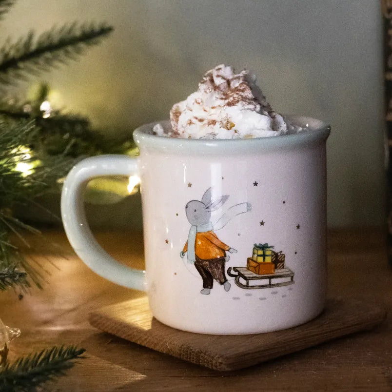 Mug with a design of a rabbit and a sleigh, filled with hot chocolate and whipped cream, on a wooden surface with a Christmas tree in the background.