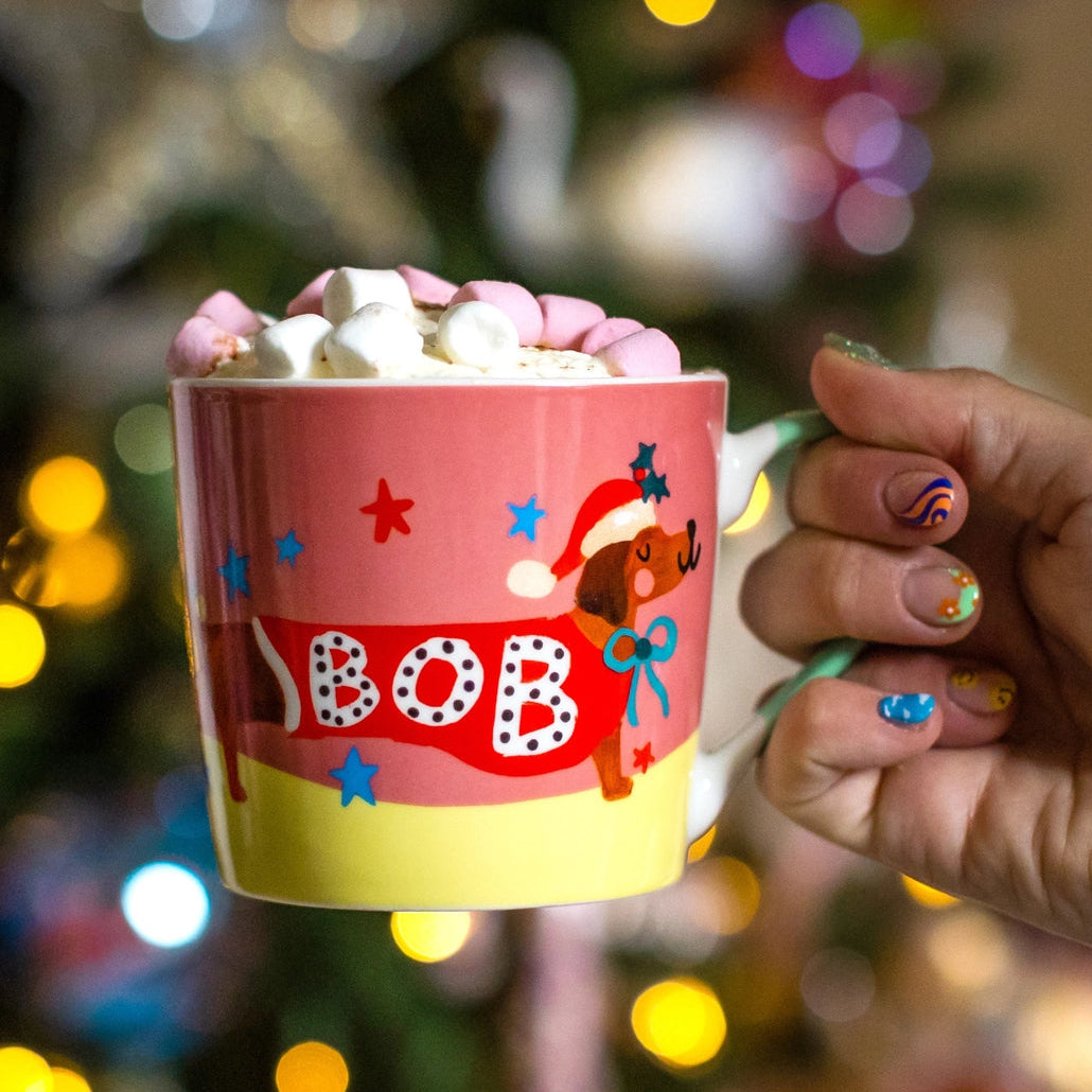 Person holding a pink mug with 'BOB' and a dog design, filled with hot chocolate and marshmallows, against a festive background.