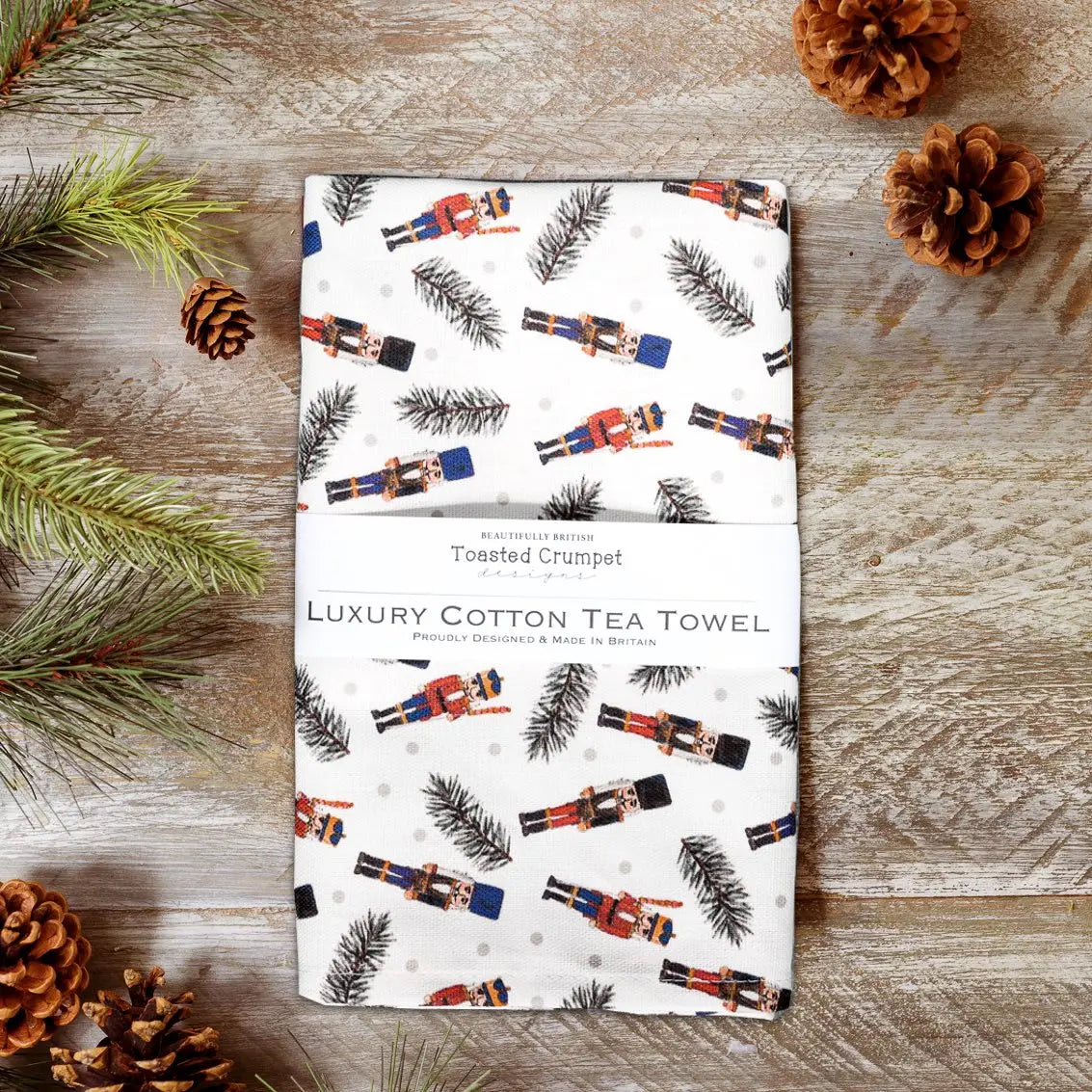 Luxury cotton tea towel with nutcracker design on a wooden surface with pine cones and branches.