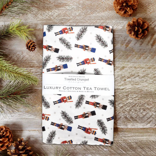 Luxury cotton tea towel with nutcracker design on a wooden surface with pine cones and branches.