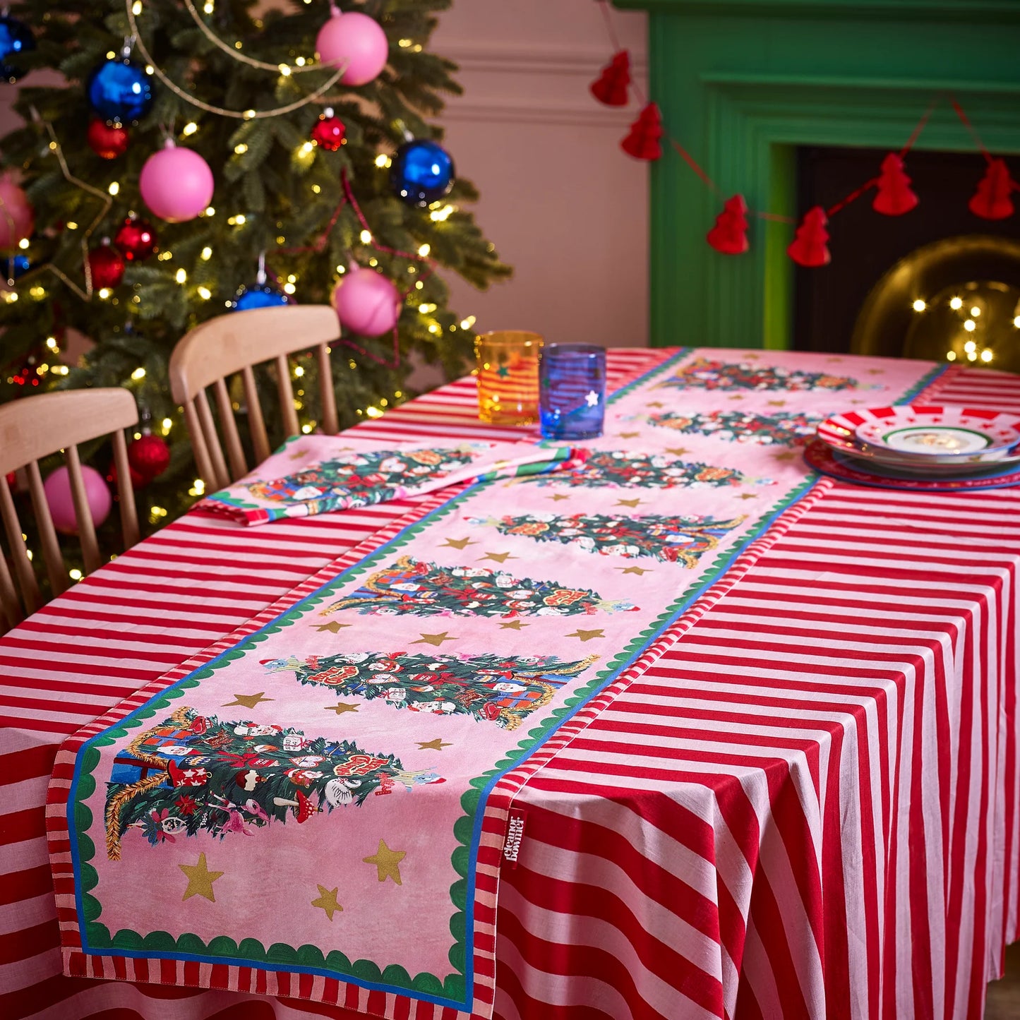 Decorative table setting with a Christmas tree-themed runner and striped tablecloth in front of a decorated Christmas tree.
