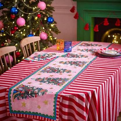 Decorative table setting with a Christmas tree-themed runner and striped tablecloth in front of a decorated Christmas tree.