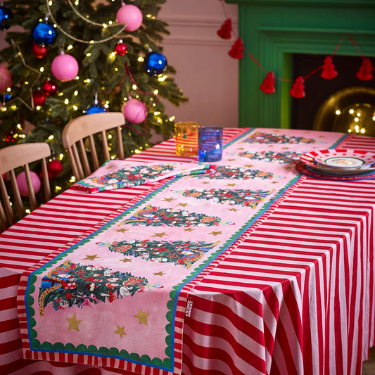 Decorative table setting with a Christmas tree-themed runner and striped tablecloth in front of a decorated Christmas tree.