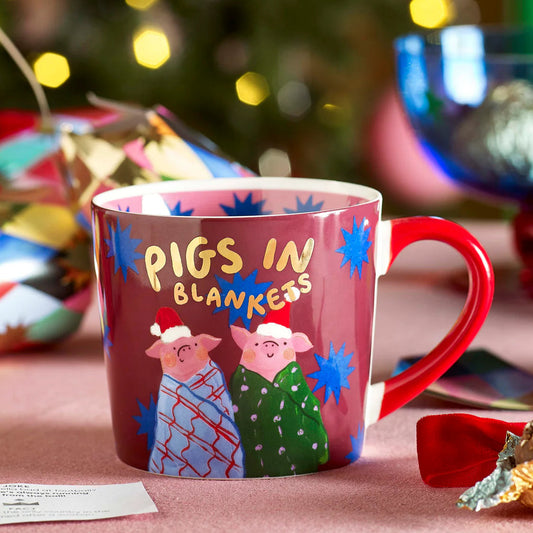 Mug with 'Pigs in Blankets' design in front of a Christmas tree with decorations.